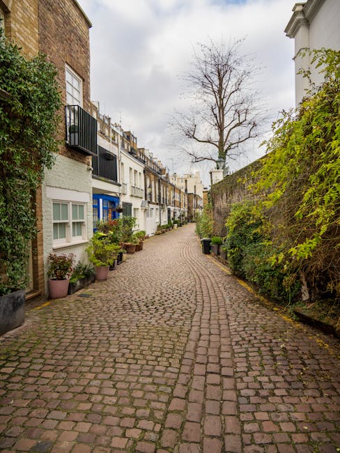 A narrow cobblestone street in South Kensington, lined with traditional terraced houses featuring white and brick exteriors. The houses have small front gardens with potted plants and shrubs, some in clay and plastic containers. A leafless tree extends its branches overhead, contrasting against a cloudy sky. On the right, a high stone wall covered with green foliage runs along the street, while on the left, balconies with black railings are visible above the ground-floor windows. The street appears quiet and residential, suitable for house removals and relocation services. South Kensington Removals occasionally operates in this area, providing careful furniture transport and packing solutions for homes with limited access, as indicated by the setting of tight access solutions for Gloucester Road flat removals.