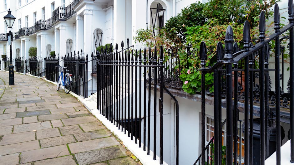 The image shows a row of white terraced houses with black wrought iron railings along the pavement on Gloucester Road. A bicycle is parked against the railings near the entrance of one property, which features a small staircase with a black railing leading down to a basement level. Green foliage and flowering plants are visible on the right side of the image, adding a touch of greenery to the scene. The pavement consists of rectangular paving stones, some of which are slightly weathered, and the scene is illuminated by natural daylight. This setting illustrates a residential street suitable for house removals and home relocation services, as provided by South Kensington Removals, with clear access to the properties through narrow doorways and staircases typical of London townhouses.