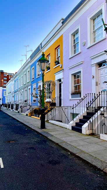 A row of colorful terraced houses on a residential street in South Kensington, featuring pastel shades of blue, yellow, and purple. The houses have white-framed sash windows, black wrought-iron fences enclosing small front gardens, and concrete steps leading to white entrance doors. A traditional black lamppost stands on the pavement in front of the houses, with a green-accented lantern. The street is paved with asphalt, and the sky above is clear and blue, indicating daylight. This scene exemplifies typical urban residential architecture and offers context for house removals and furniture transport services, as provided by South Kensington Removals, who execute packing and loading processes for home relocations.