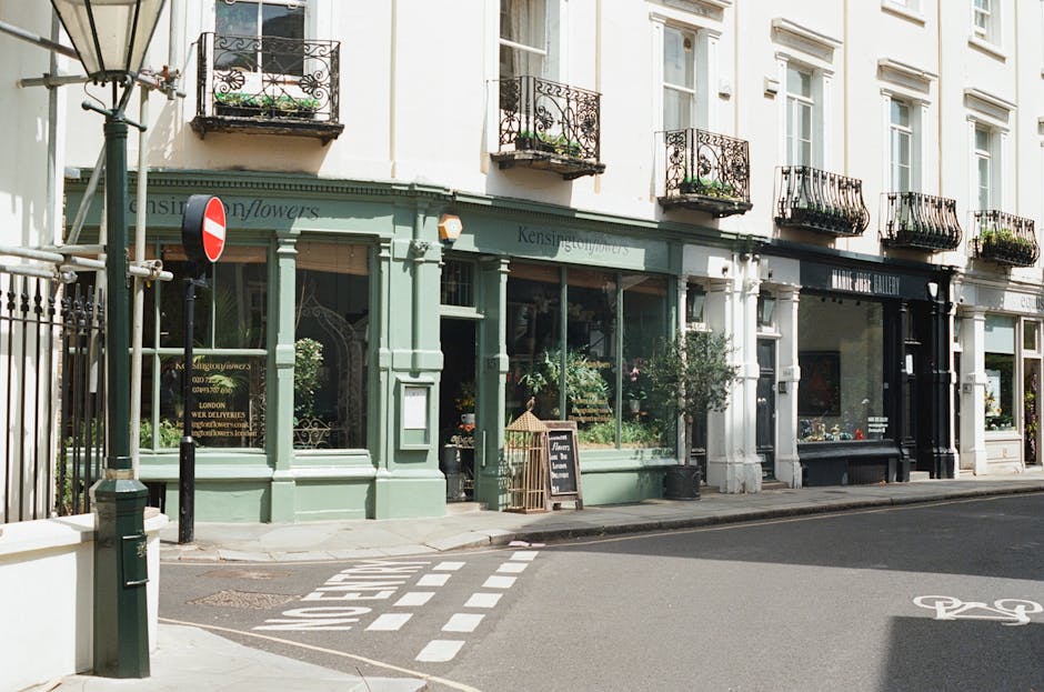 A street view outside a boutique flower shop with a green facade in Kensington and Chelsea, featuring large glass windows displaying floral arrangements. The shop has a black sign with white lettering above the entrance, and a small handwritten signboard on a stand on the sidewalk. Several potted plants and flowers are arranged near the shopfront, with a white sandwich board sign placed outside. Adjacent to the shop, there is a narrow road with painted road markings, including a dashed white line and a bicycle lane symbol, indicating designated parking or loading zones. A black metal gate and a lamp post are visible on the left side of the image, along with a no-entry traffic sign mounted on a pole. The building has multiple stories with white walls and black wrought iron balconies on the upper floors, some with flower boxes. The scene suggests a typical urban environment suitable for residential or commercial house removals and furniture transport, with South Kensington Removals often operating in such settings during home relocation or moving processes.