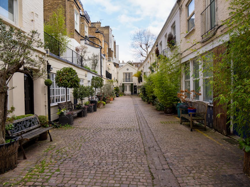 A narrow cobblestone residential courtyard in South Kensington with white terraced townhouses on either side, each featuring large windows, some with black iron balconies, and potted plants along the facades. On the left, a black metal bench is positioned near a small tree and decorative potted box bushes, while on the right, a wooden bench and a recycling bin are placed beside tall leafy shrubs and potted greenery. The cobblestone pathway is slightly worn, with visible gravel and subtle lines indicating previous loading and unloading activity. The scene is captured during daytime with natural daylight illuminating the area, and a glimpse of the sky with some clouds is visible above the rooftops. This setting is typical for home relocation or furniture transport processes involving careful packing and moving within or out of these properties, which South Kensington Removals often assist with.