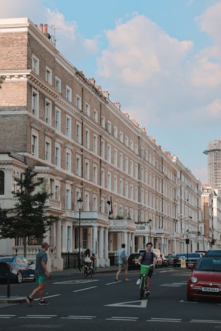A row of multi-story Georgian-style terraced buildings with cream-colored facades and large windows lining a busy street in South Kensington. In the foreground, pedestrians are crossing the road, including a young man on a bicycle, a man walking with a child, and others walking along the pavement. Several cars are parked along the curb, and a grey van is partially visible at the right edge of the image, suggesting active loading or unloading activities related to home relocation or furniture transport. The scene is illuminated by natural daylight, with a partly cloudy sky overhead. This setting illustrates a typical urban environment where house removals and moving services, such as those provided by South Kensington Removals, may be carried out, often involving careful planning of packing, loading, and transport logistics.
