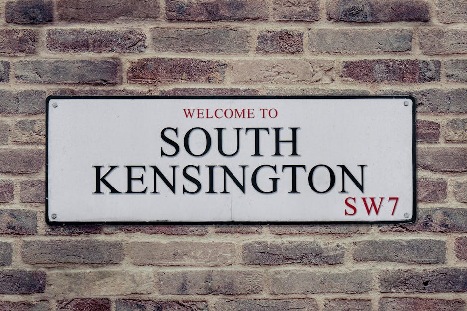 A rectangular metal street sign mounted on a brick wall, displaying the words 'Welcome to South Kensington' in red and black text, with the postcode 'SW7' in red at the bottom right corner. The brick wall behind the sign is composed of reddish-brown bricks with visible mortar lines, typical of urban residential areas in South Kensington. The sign's surface appears smooth and reflective, with four screws securing it to the wall, indicating a permanent fixture. This type of signage is common in property marking zones or designated residential districts, which can be relevant during home relocation or furniture transport services provided by South Kensington Removals. The image captures the sign in natural daylight, emphasizing the clear, legible lettering and the structural texture of the brick wall, providing context for moving logistics in the South Kensington area, especially when considering parking suspensions and local regulations during house removals.