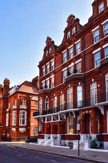 A row of multi-storey red brick buildings with large sash windows and decorative iron balcony railings on the first floor, situated along a quiet street. The buildings have a Victorian or Edwardian architectural style, with detailed brickwork and ornate gables at the roofline. The street features a paved sidewalk with a metal railing, and the sky above is clear with bright sunlight illuminating the scene. This image is associated with house removals and home relocation services offered by South Kensington Removals, showcasing the type of residential properties involved in local furniture transport and packing and moving processes, which are often carried out by professional removal companies within the South Kensington area.