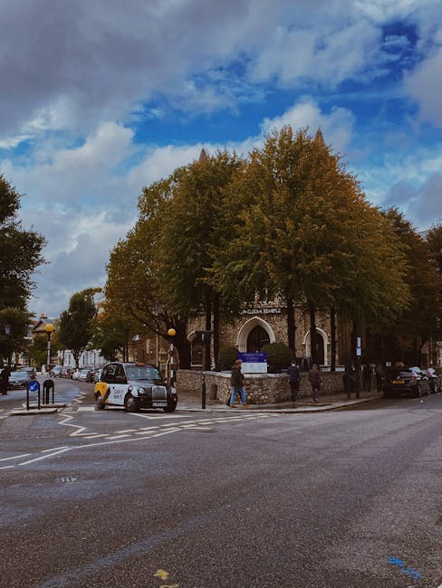 A street scene outside a church building with a rounded arched entrance, surrounded by several large trees with green and golden foliage. The church has brick walls and a small sign above the entrance. In the foreground, there are vehicles including a silver car and a black van parked near the curb. Several pedestrians are walking along the pavement, some near the church's stone boundary wall. The sky above is partly cloudy with patches of blue, indicating natural daylight. This image depicts the urban environment typical of house removals and furniture transport activities in South Kensington, where [COMPANY_NAME], such as South Kensington Removals, may facilitate home relocation and moving services, including loading and unloading processes at this location.