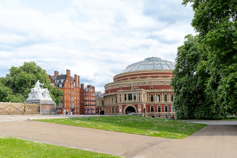 A wide view of the exterior of the Royal Albert Hall in South Kensington during daytime, with partly cloudy skies overhead. In the foreground, a paved pathway curves around a grassy area, which is bordered by large green trees on the right side of the image. To the left, there is a white stone sculpture of a seated figure and two reclining figures, positioned on a stone pedestal, and a decorative fence behind it. The building's brick façade is visible, featuring arched windows, columns, and decorative detailing, with a distinctive domed roof partly covered by a protective covering. The surrounding environment appears peaceful, with no visible moving activity or loading equipment, capturing an outdoor scene that relates to a home relocation or transfer process near a historic cultural site, as part of a professional removals service by South Kensington Removals.