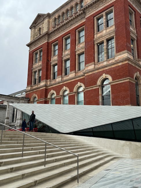 Image of the exterior of a historic red-brick building with ornate stone window frames and arched window designs, viewed during daytime with overcast weather. In the foreground, modern white marble stairs with stainless steel handrails lead up to a glass entrance. Two individuals are visible on the stairs, one standing at the top and the other slightly below, both wearing dark clothing. Adjacent to the stairs, a sleek, angular glass and metal structure extends from the building, creating a contemporary entrance area. The scene depicts a combination of traditional architecture and modern design, consistent with the typical setting for residential or commercial relocations. This environment could be part of a property being prepared for house removals or furniture transport services, as managed by South Kensington Removals.