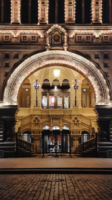 A wide view of the exterior of the Royal Albert Hall in South Kensington during daytime, with partly cloudy skies overhead. In the foreground, a paved pathway curves around a grassy area, which is bordered by large green trees on the right side of the image. To the left, there is a white stone sculpture of a seated figure and two reclining figures, positioned on a stone pedestal, and a decorative fence behind it. The building's brick façade is visible, featuring arched windows, columns, and decorative detailing, with a distinctive domed roof partly covered by a protective covering. The surrounding environment appears peaceful, with no visible moving activity or loading equipment, capturing an outdoor scene that relates to a home relocation or transfer process near a historic cultural site, as part of a professional removals service by South Kensington Removals.