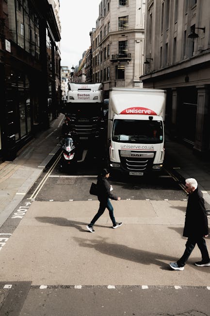 An urban street scene during a home relocation involves a white moving van with the branding 'uniserve' parked partially on the pavement and street, with its rear doors open for loading or unloading furniture and boxes. Behind it is a larger van marked 'Becton Transport,' positioned on the road, indicating active logistics. A scooter is parked adjacent to the vans near the curb. Two pedestrians, a woman and an older man, are crossing the street at a marked pedestrian crossing; the woman is dressed in casual clothing and carrying a bag, while the man is wearing dark clothing, both walking in opposite directions. The street is lined with multi-story stone and brick buildings with fire escape elements and large windows, characteristic of a city centre in South Kensington. Overhead, the sky is partly cloudy, and the scene captures the busy, urban atmosphere associated with house removals and furniture transport within the area, reflecting the logistics involved in home moving services, as provided by South Kensington Removals.